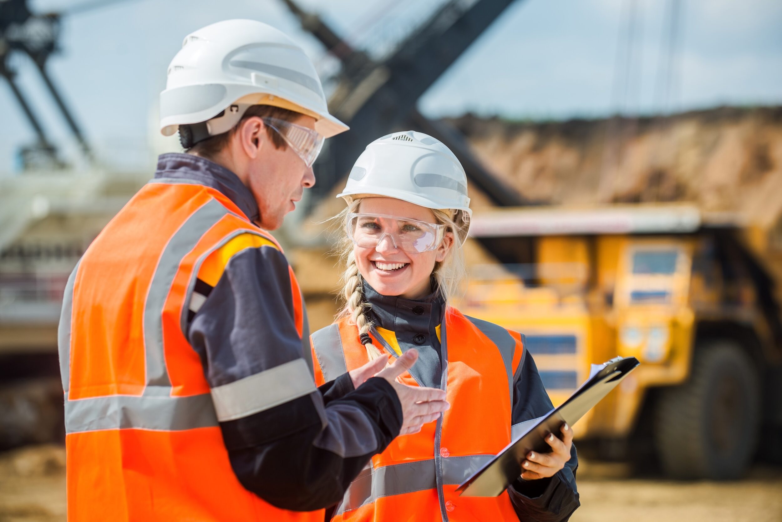 Two workers wearing hard hats and safety vests talking while looking at a clipboard at a mining site