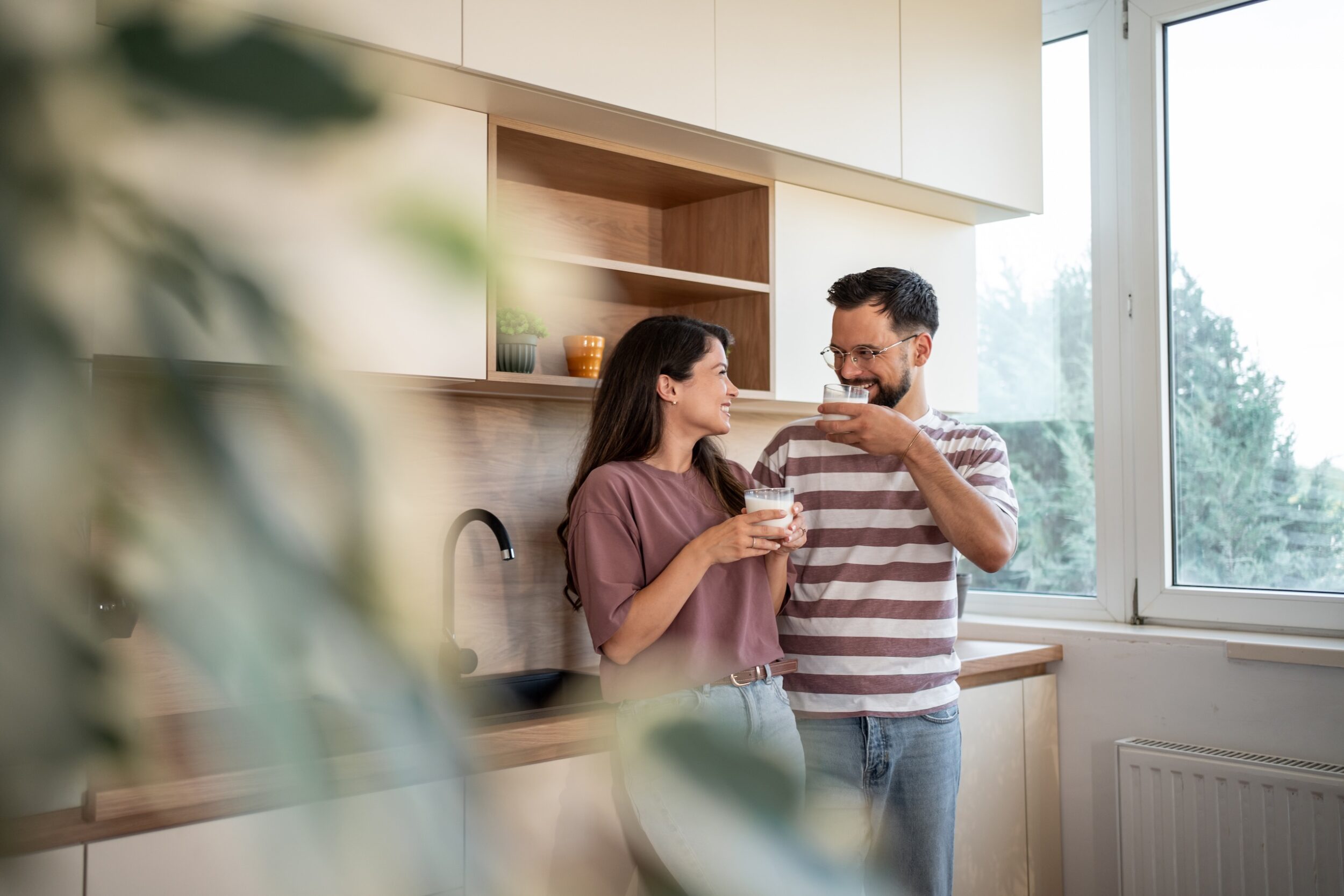 Young couple is standing in their modern kitchen, holding glasses of milk and smiling at each other, enjoying a quiet moment together before starting their day