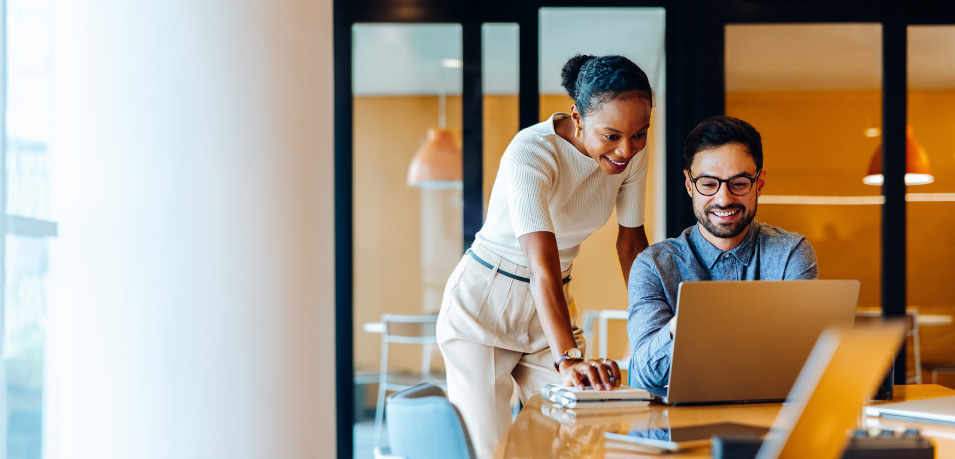 Woman in white top and man in blue shirt working together on laptop at desk in modern glass office