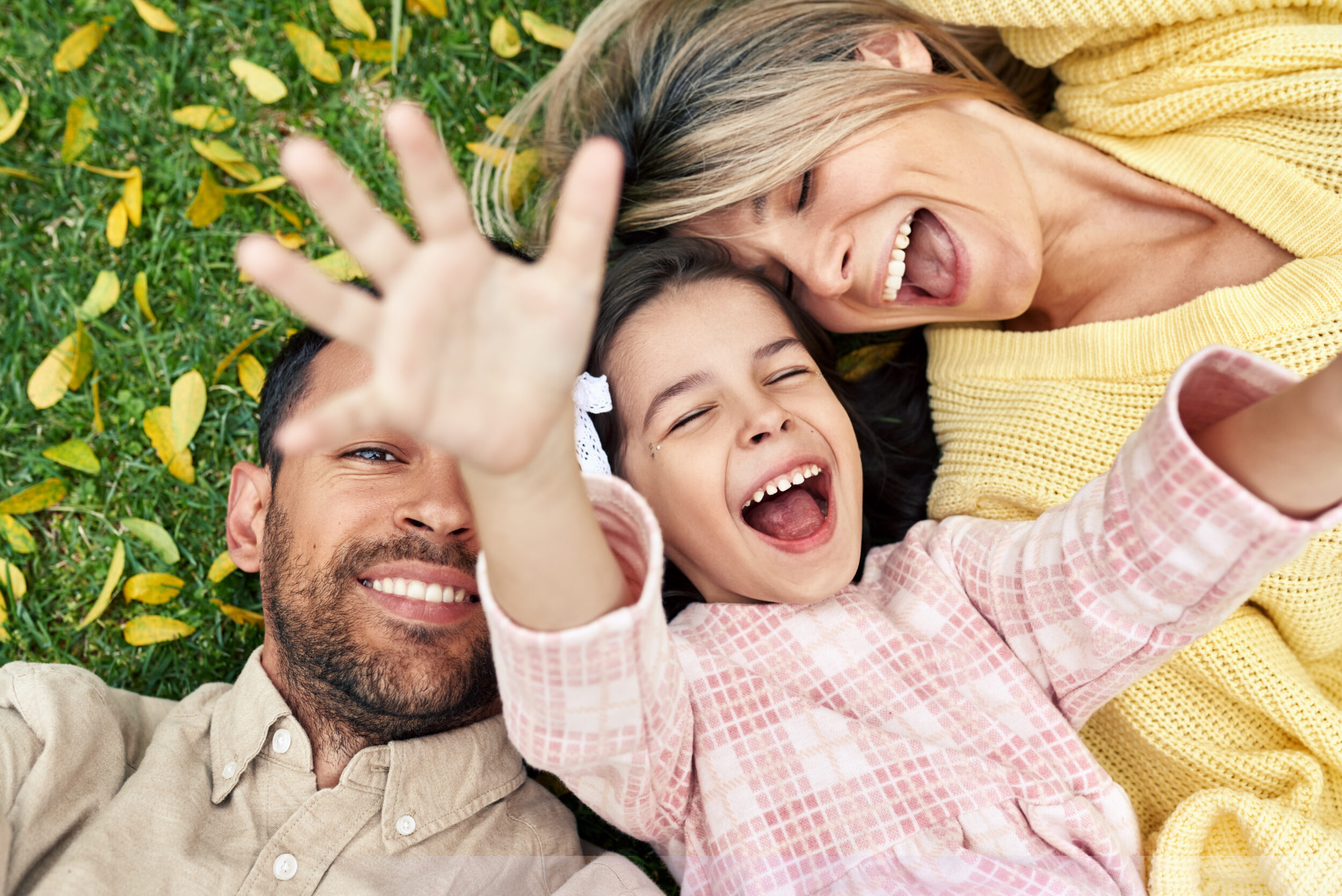 Two adults and a child lying on grass outdoors, smiling while the child reaches toward the camera