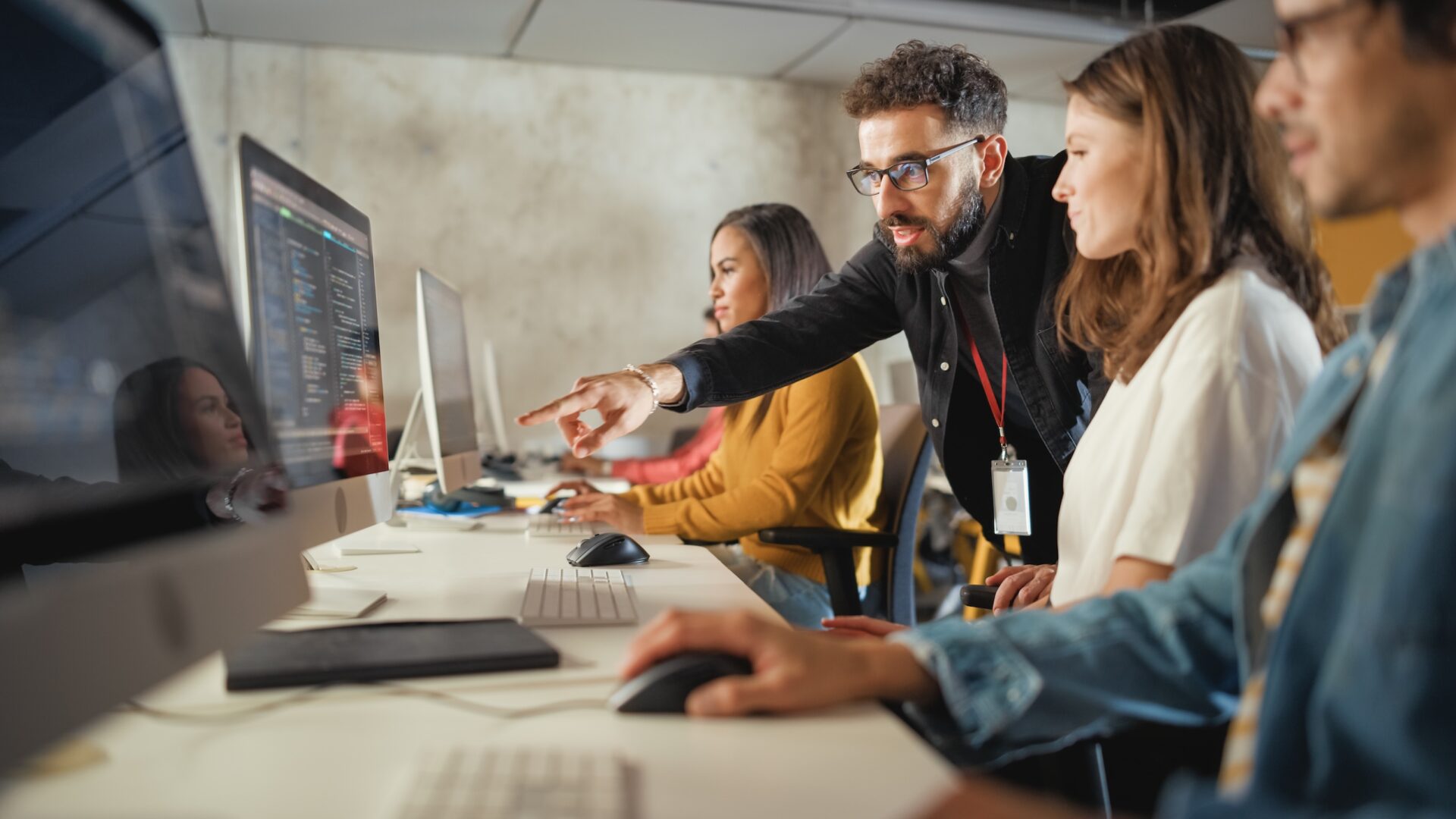 Man in black shirt pointing at computer screen while collaborating with colleagues at desk workstation