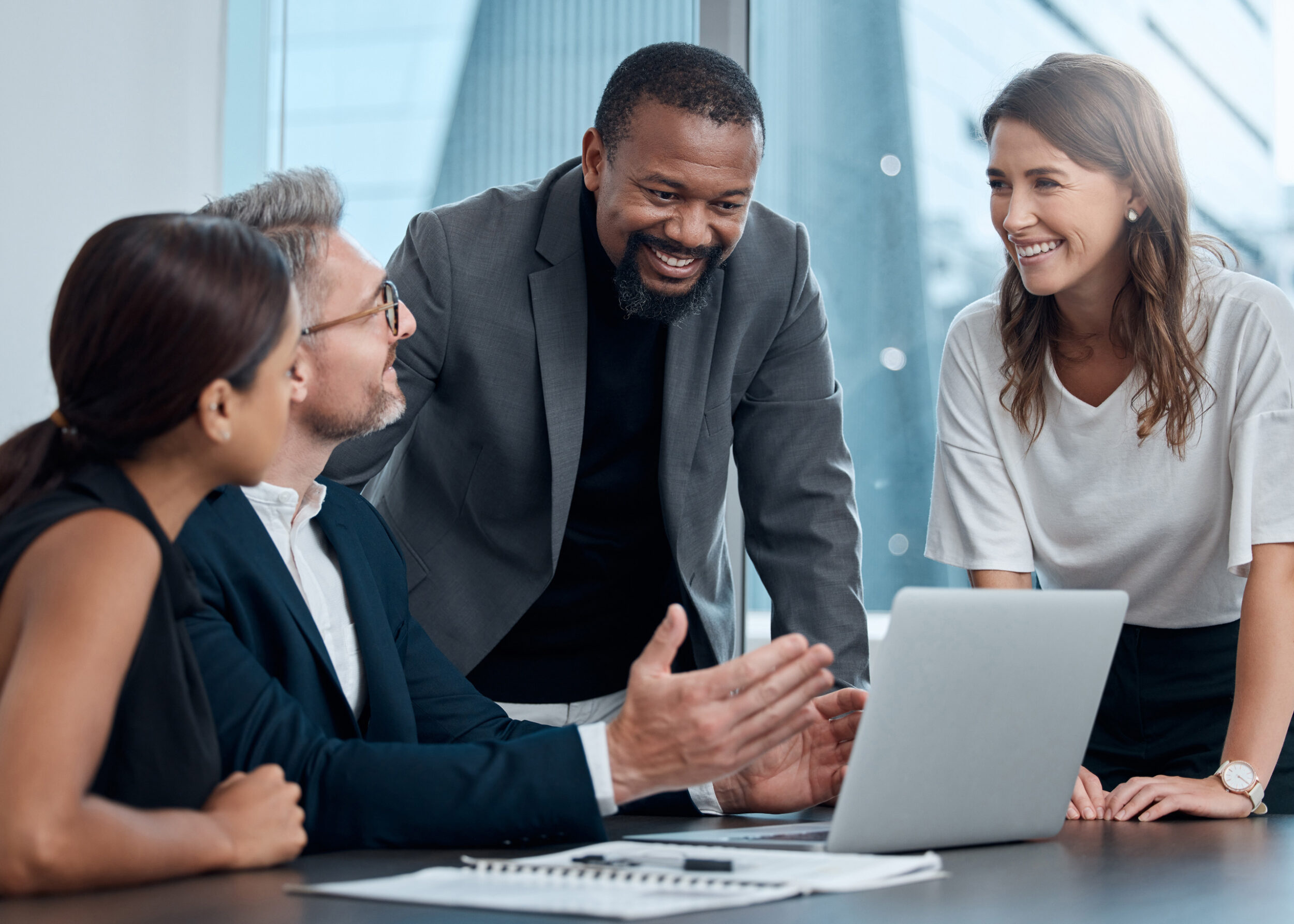 Four professionals gathered around laptop at boardroom table in modern glass-walled office building