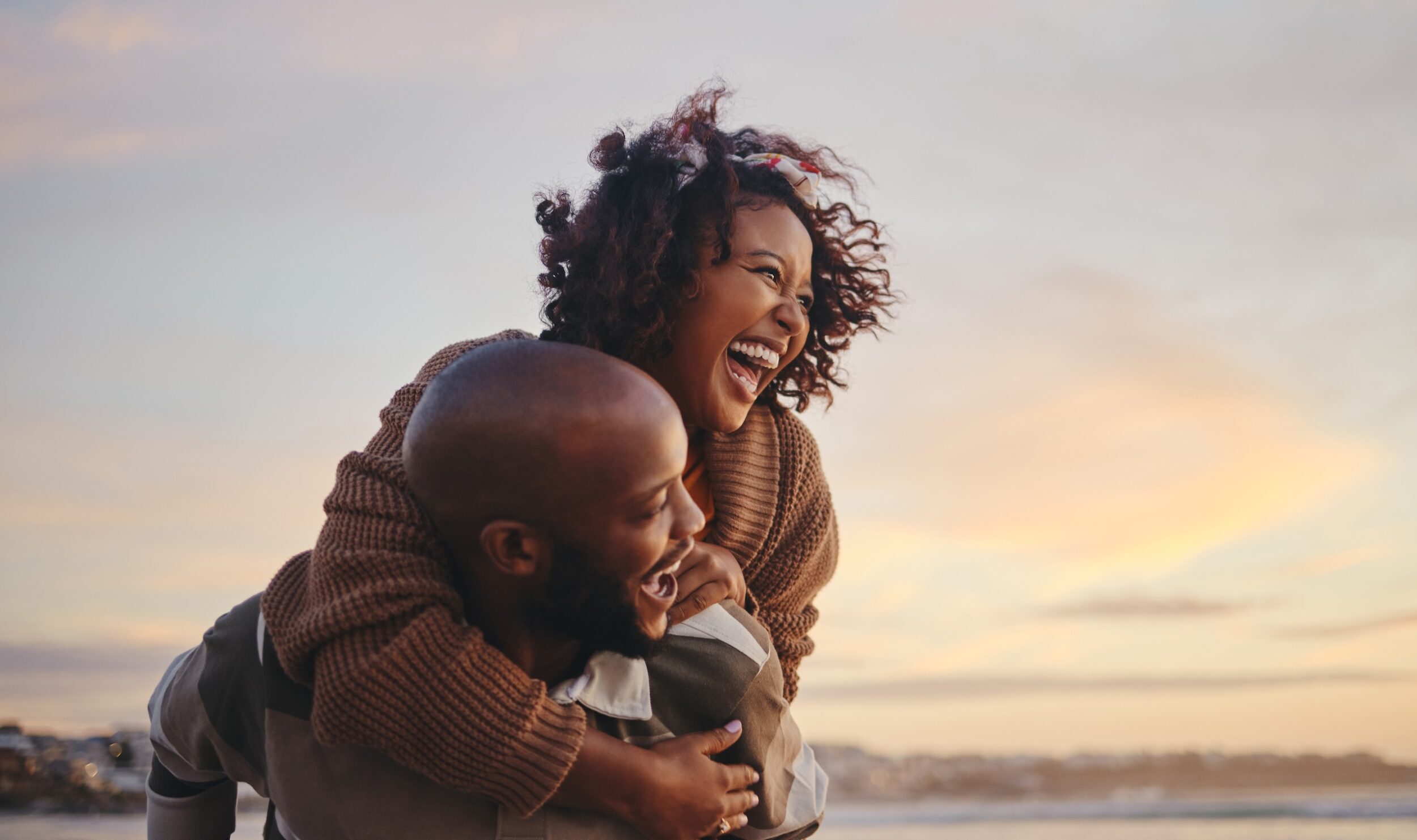 Couple laughing together outdoors near the coast, with one person carrying the other on their back