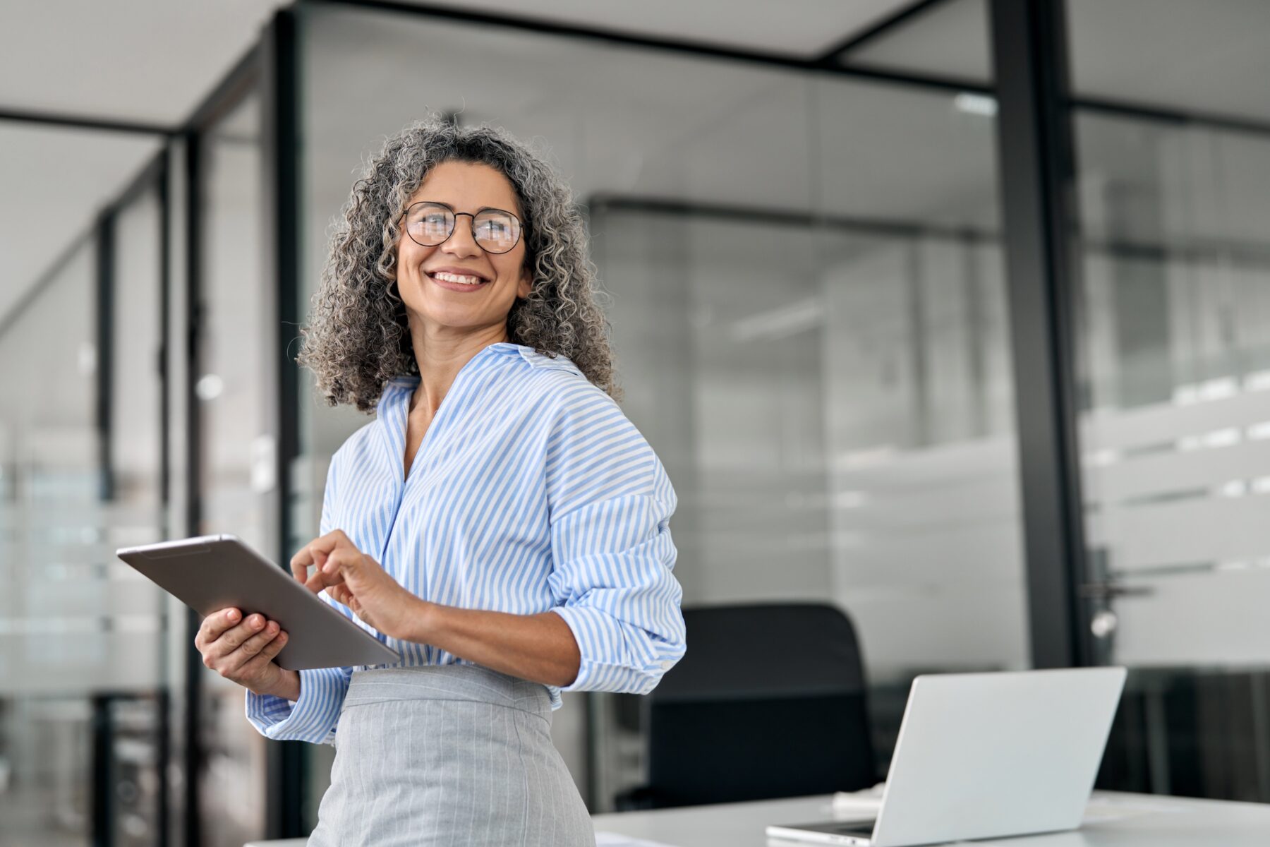 Woman standing in a modern office holding a tablet, smiling while looking slightly to the side