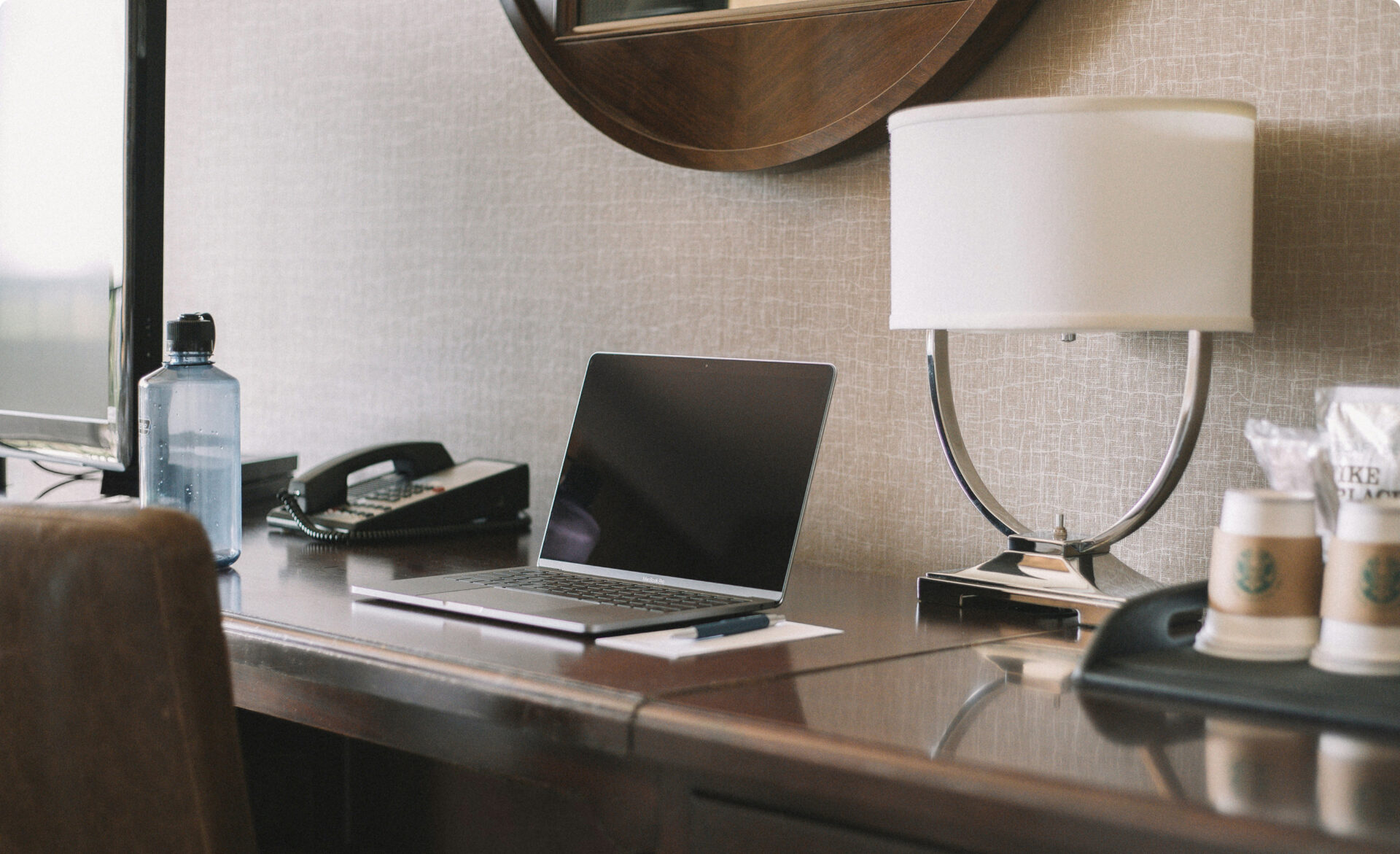 Laptop placed on a desk beside a lamp, phone, water bottle and cups in a hotel-style room