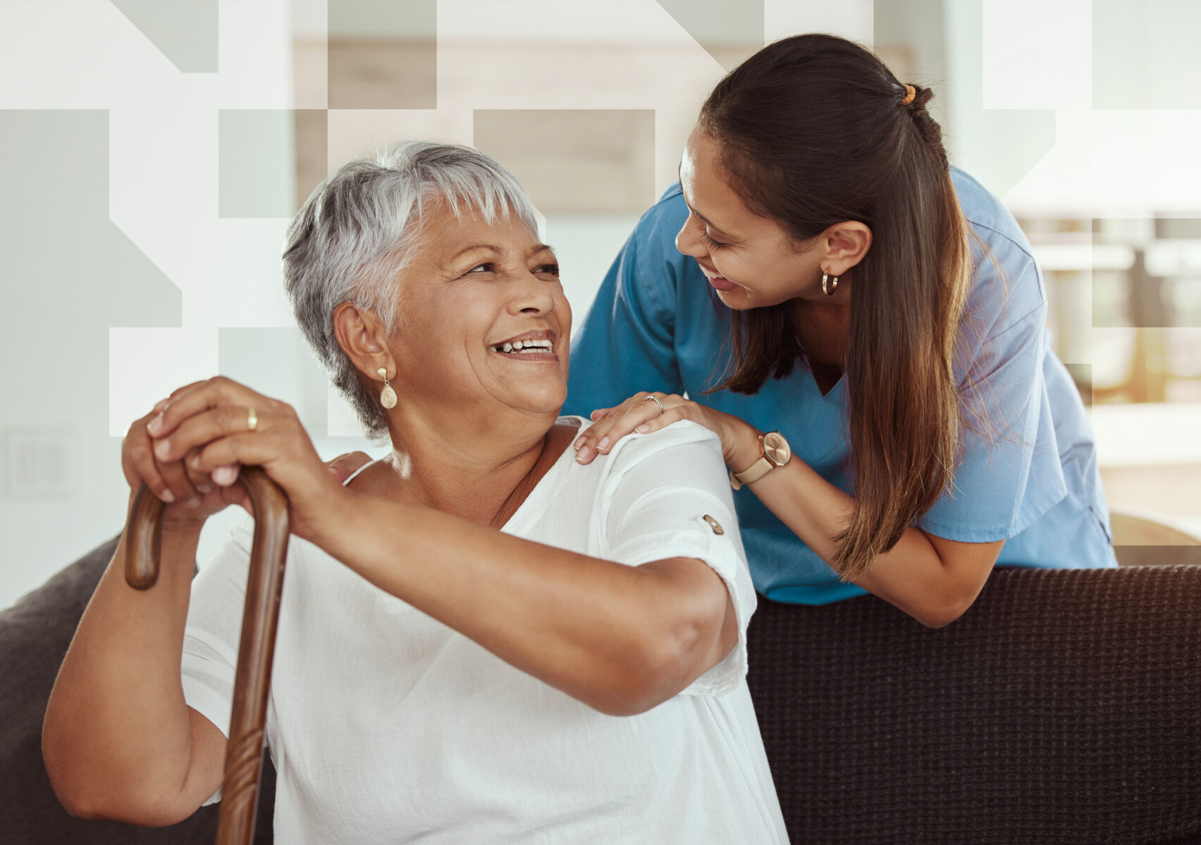 Care worker leaning toward an older woman seated indoors, smiling while resting a hand on her shoulder