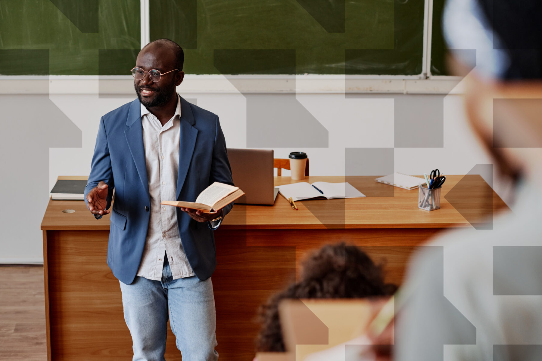 Person standing at the front of a classroom holding a book while speaking to people seated nearby